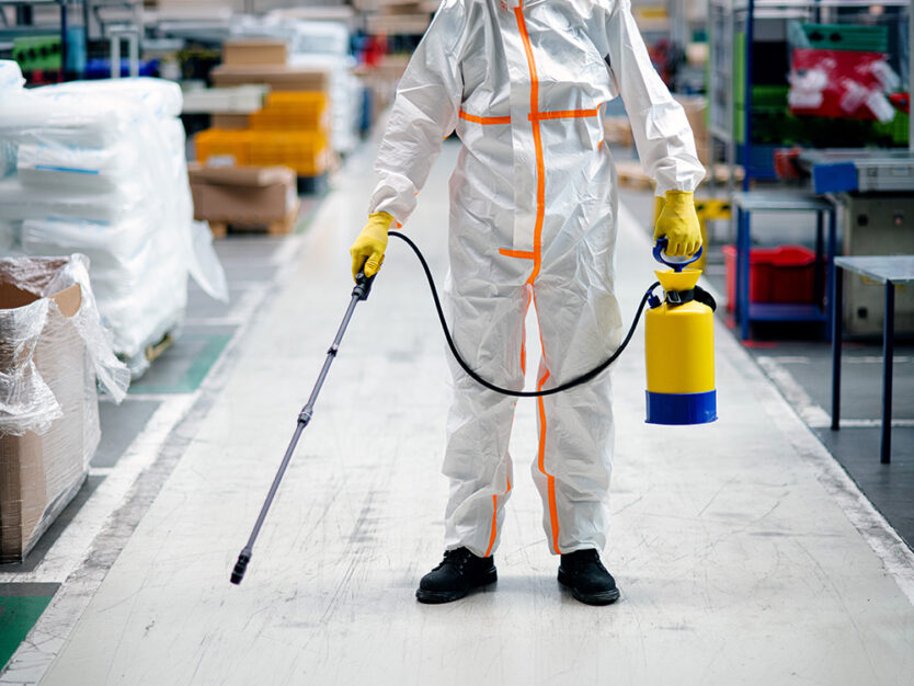 Front view portrait of man worker with protective mask and suit disinfecting industrial factory with spray gun.
