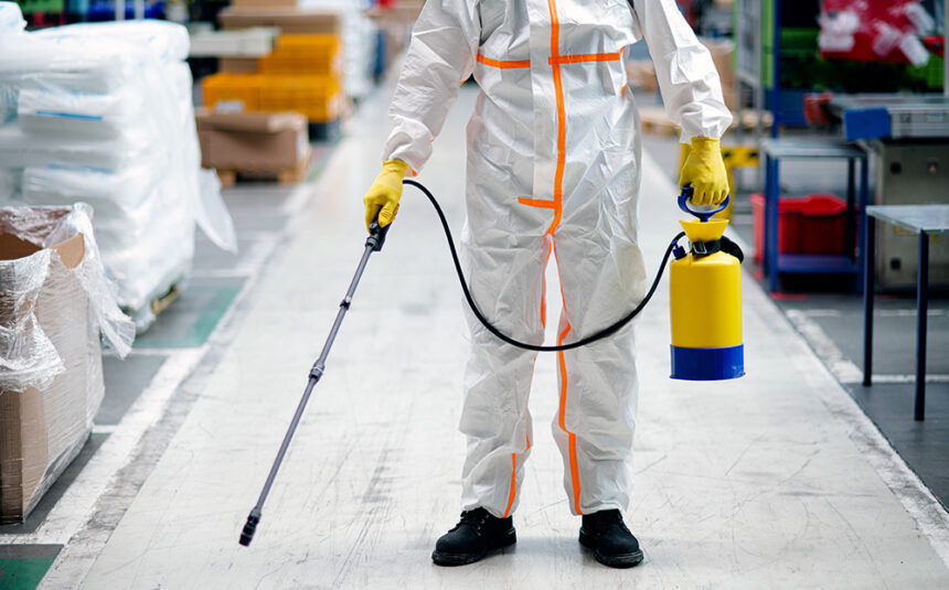 Front view portrait of man worker with protective mask and suit disinfecting industrial factory with spray gun.
