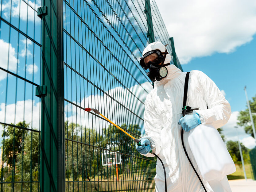 male specialist in hazmat suit and respirator disinfecting fence of basketball court in park during