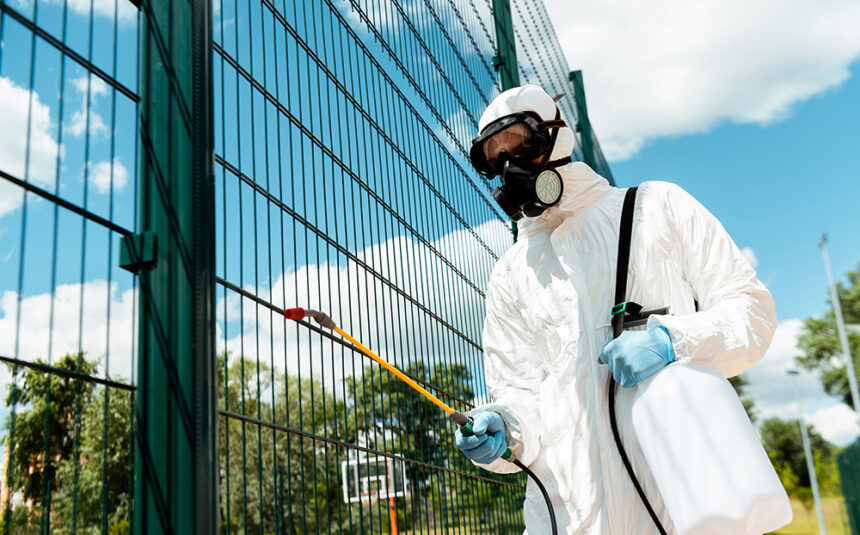 male specialist in hazmat suit and respirator disinfecting fence of basketball court in park during