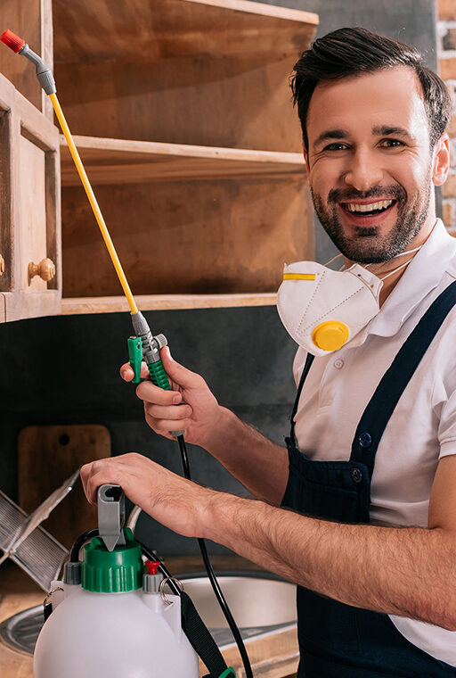 smiling pest control worker spraying pesticides on shelves in kitchen