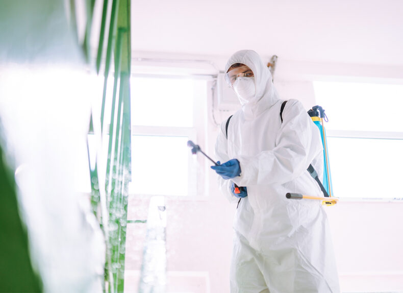 Man in Hazmat suit, protective gloves and goggles use sprayer equipment disinfect the entrance.