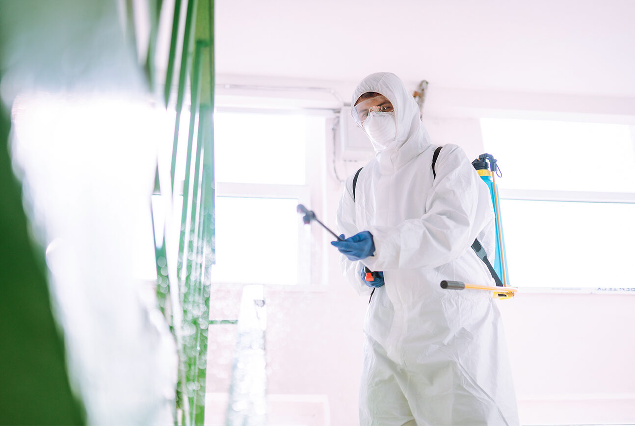 Man in Hazmat suit, protective gloves and goggles use sprayer equipment disinfect the entrance.